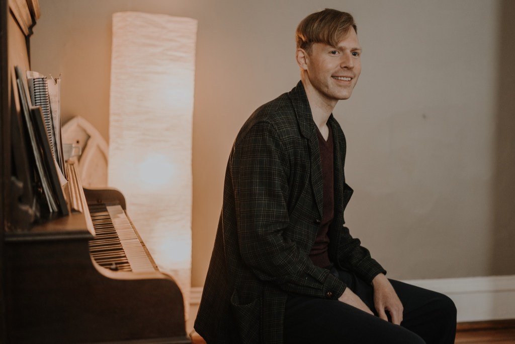 A smiling man sitting beside a piano, wearing a plaid jacket and a maroon shirt, with a warm lamp in the background.