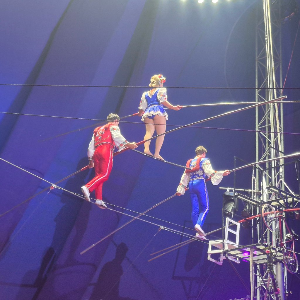 Three performers walking on a tightrope during Circus Flora, showcasing a colorful display of costumes under the tent's bright lights.