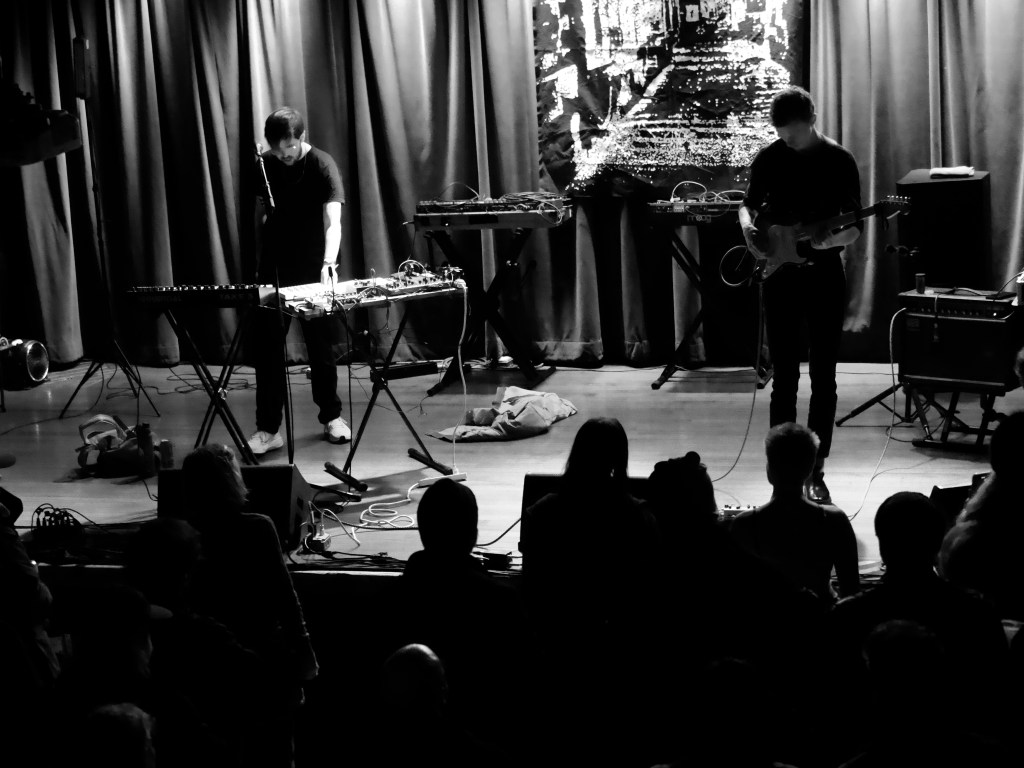 Two musicians performing on stage with electronic instruments in a dimly lit venue. The audience is visible, engaged in the performance.