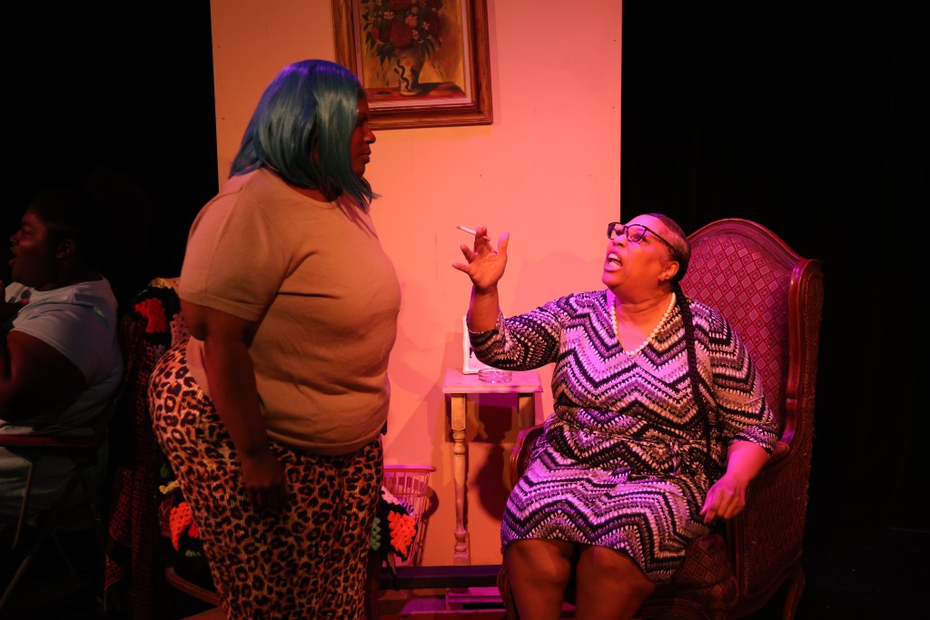 A scene from the play _Heavy_, featuring two women in a heated discussion, with one sitting in a patterned dress and glasses, and the other standing with blue hair and a tank top. The setting includes a chair and a table with a flower painting on the wall.