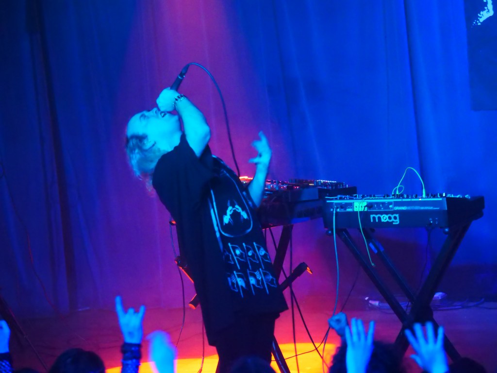 A performer singing passionately on stage with a synthesizer in the background and audience hands raised in excitement.