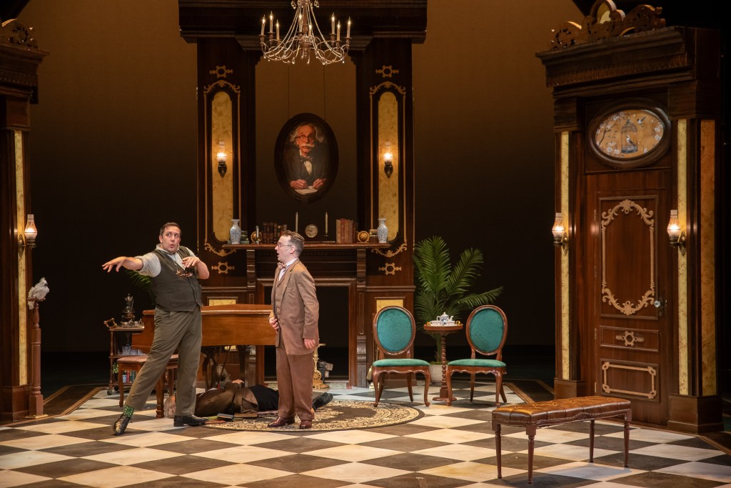 A scene from the musical mystery 'Murder for Two', featuring two male characters in a theatrical set with a chandelier, piano, and period-style furniture.