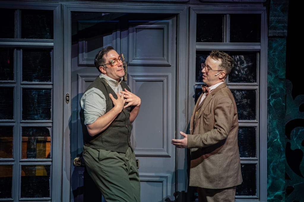 Two male actors perform in a scene from the musical 'Murder for Two,' wearing comedic expressions and costumes, set against a backdrop of a door and large windows.