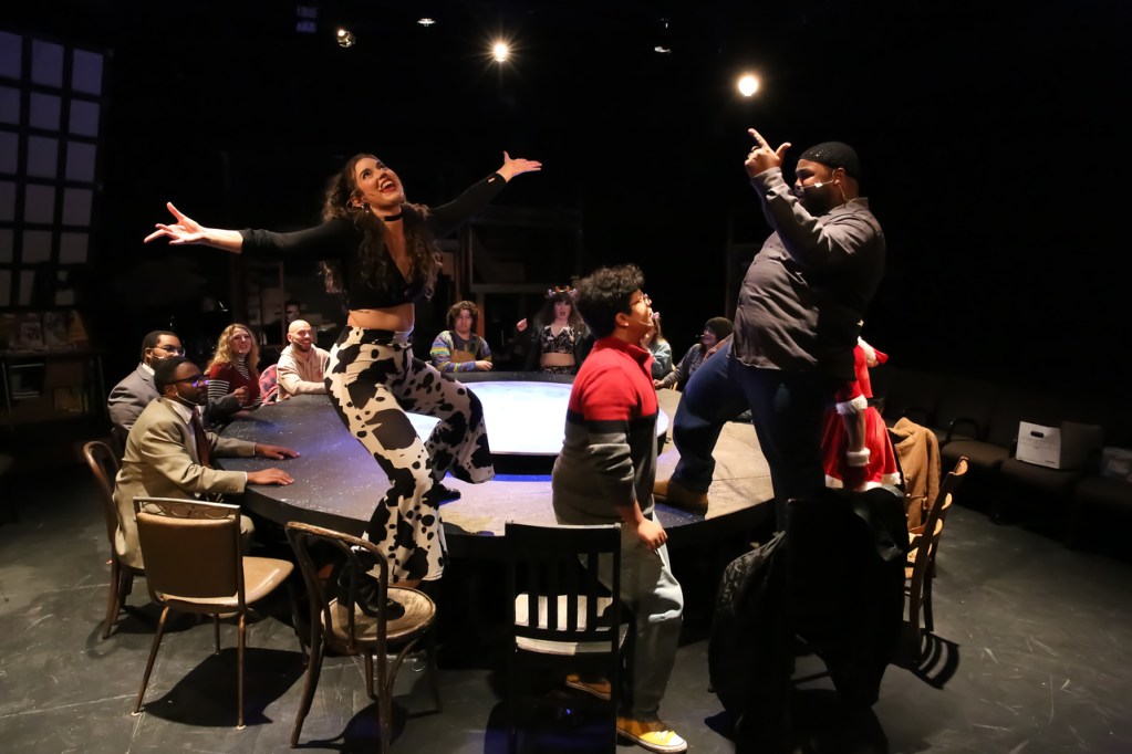 A lively scene from a theater production featuring actors engaged in a dynamic performance, with one performer striking a dramatic pose on a circular stage while the audience observes enthusiastically.