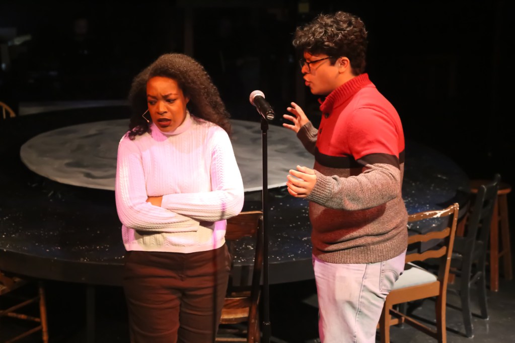 A scene from the musical Rent featuring two characters, one woman looking upset and crossing her arms, and a man passionately singing near a microphone on stage.