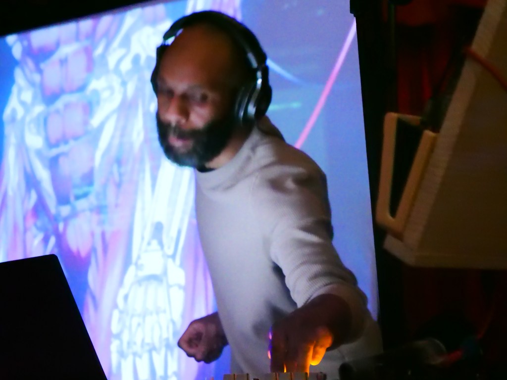 A DJ performing on stage with headphones, focused on the turntables, against a colorful backdrop.