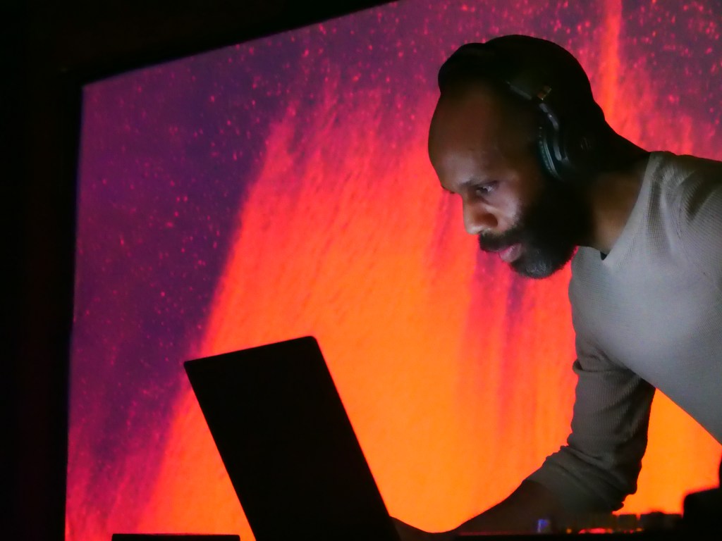 A DJ performing live in front of a vibrant, colorful backdrop while focused on his laptop.