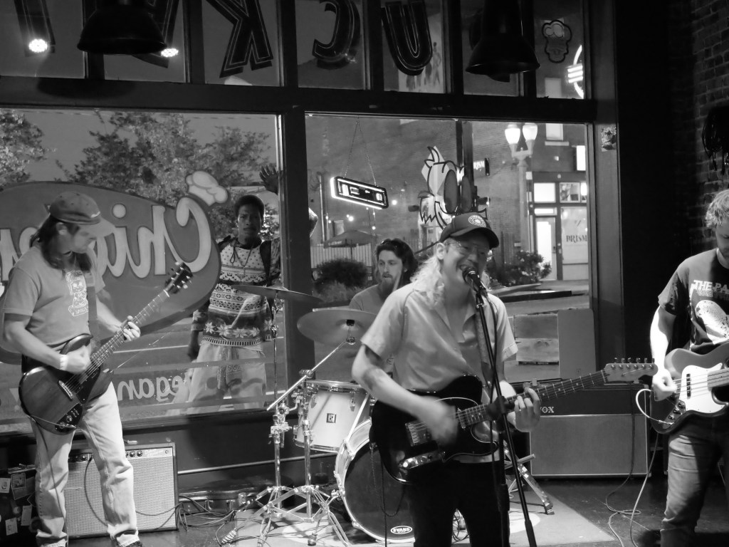 A black and white image of a live band performing inside a venue, featuring a singer with a guitar, a drummer, and a bassist, with audience members visible outside the window.