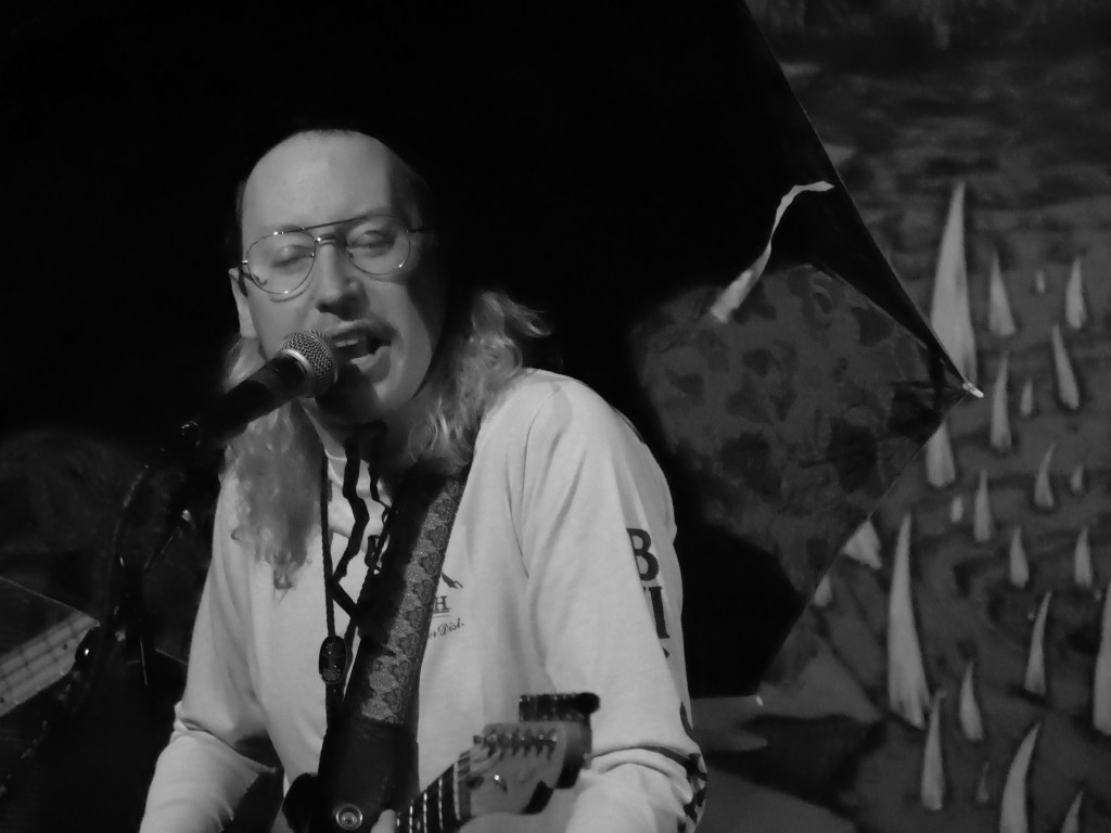 Musician performing with a microphone and guitar, captured in black and white.