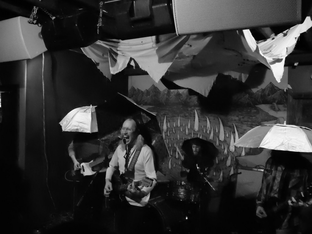 A black and white photo of a live music performance featuring a vocalist playing guitar and singing, surrounded by band members using umbrellas. The background includes artistic decorations resembling mountains and sailing boats.