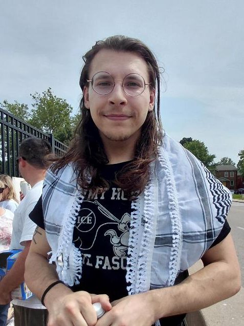 A young person with long hair and glasses wearing a black t-shirt and a patterned scarf stands outdoors during a protest.