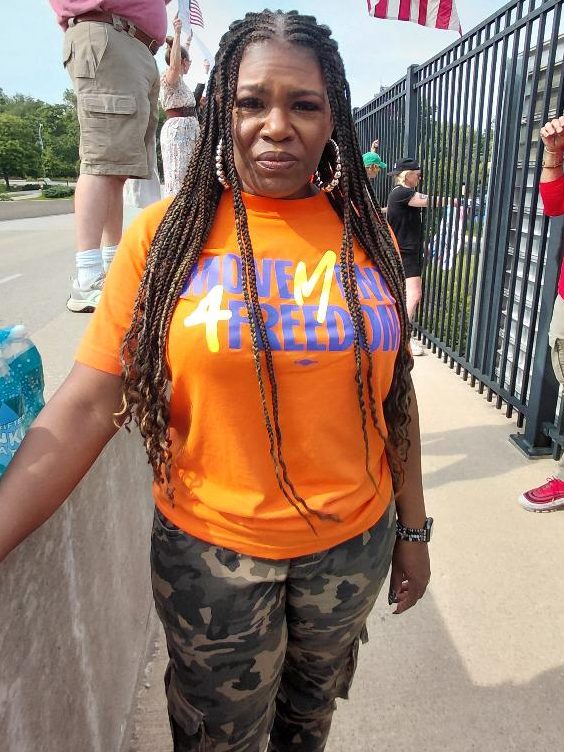 A woman with long braids wearing an orange shirt with the text 'Movement 4 Freedom' stands by a railing at an outdoor protest, with a group of people visible in the background.