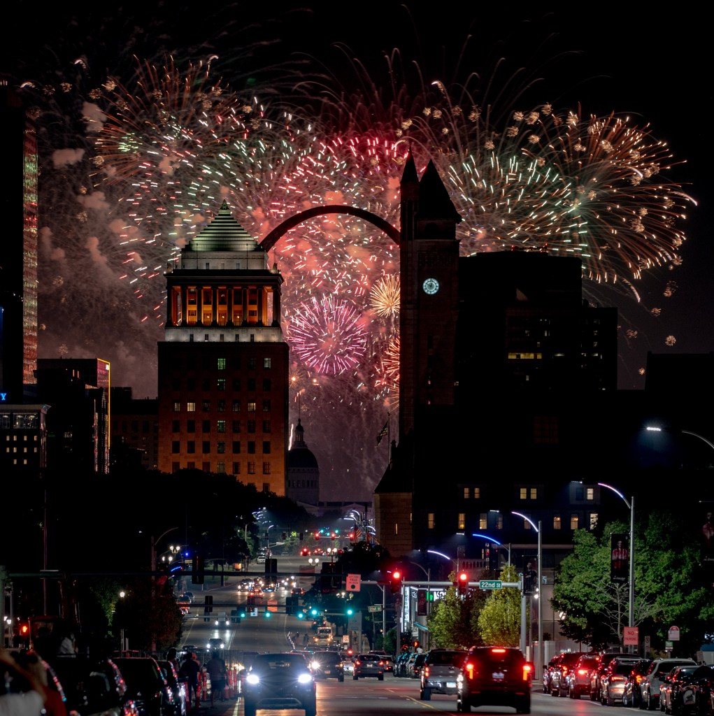 Fireworks display over the St. Louis skyline with the Gateway Arch in the background, view from a city street illuminated by traffic lights.