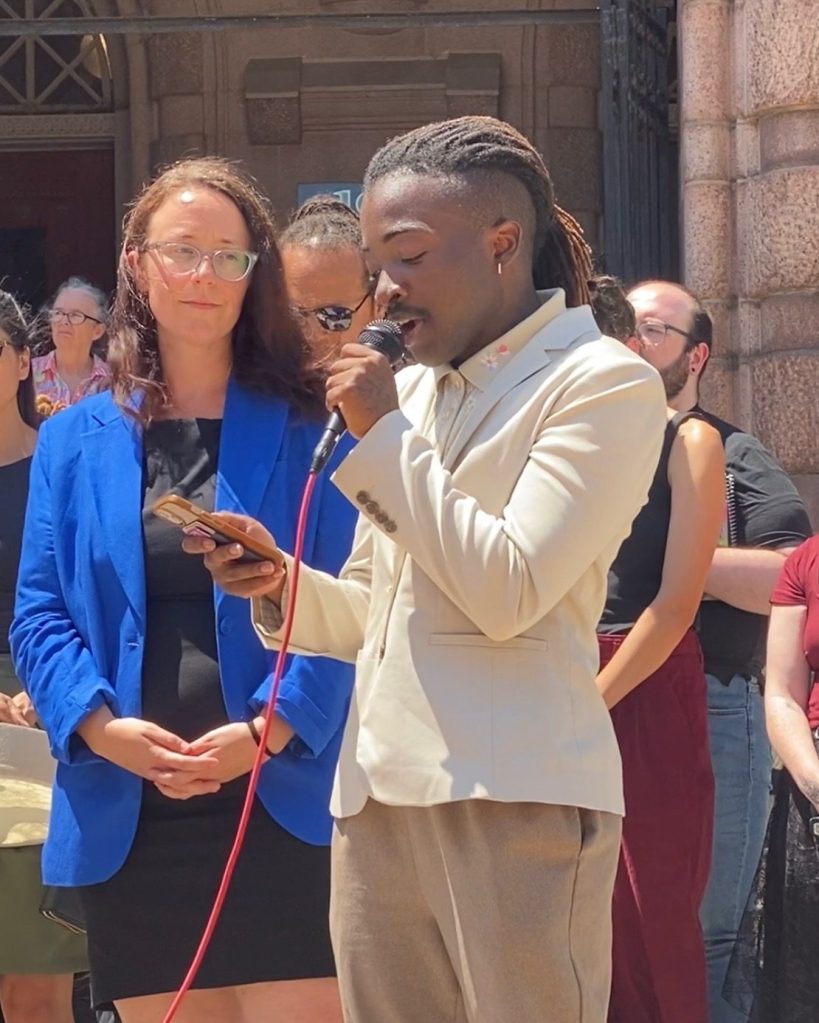 A person in formal attire speaks into a microphone during a press conference or public gathering, while an audience listens in the background. The setting appears to be outside in front of a historical building.