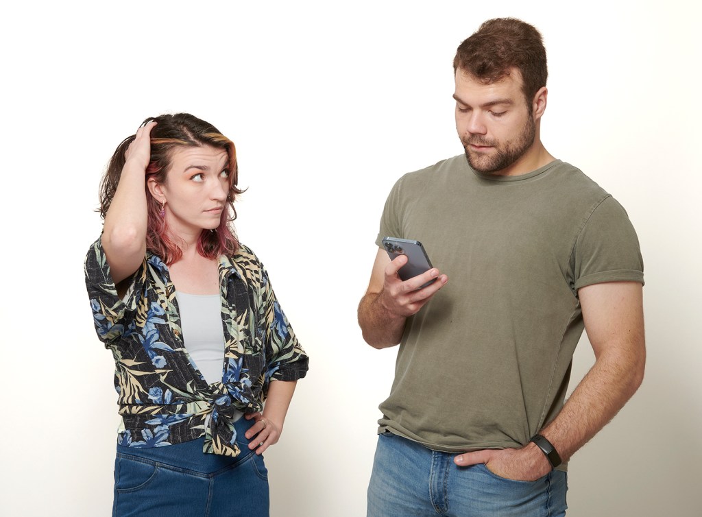 A woman with pink highlights in her hair looks concerned while tugging at her hair, standing beside a man in a gray shirt who is focused on his phone. Both are set against a plain white background.