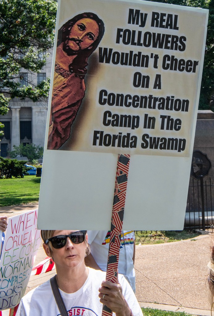 A woman wearing sunglasses holds a protest sign featuring a depiction of Jesus and a message criticizing concentration camps in Florida.