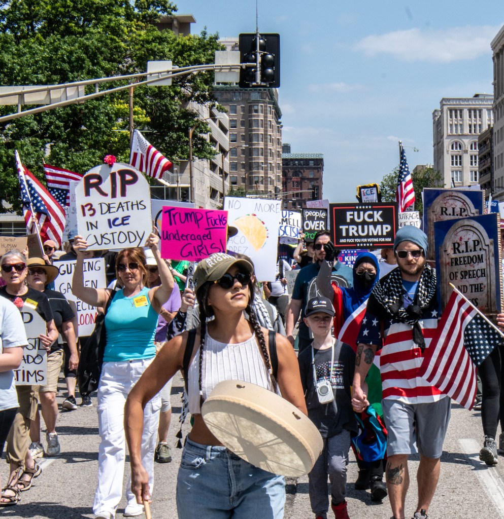 A diverse group of protesters marching in downtown St. Louis, carrying signs related to immigration issues and political dissent. Some participants are holding American flags and a drum, while others express solidarity with victims of the deportation campaign.