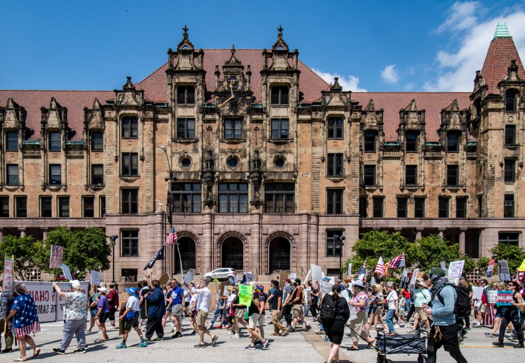 A large group of protesters marching in front of City Hall in downtown St. Louis, carrying various signs and banners, on a sunny day.