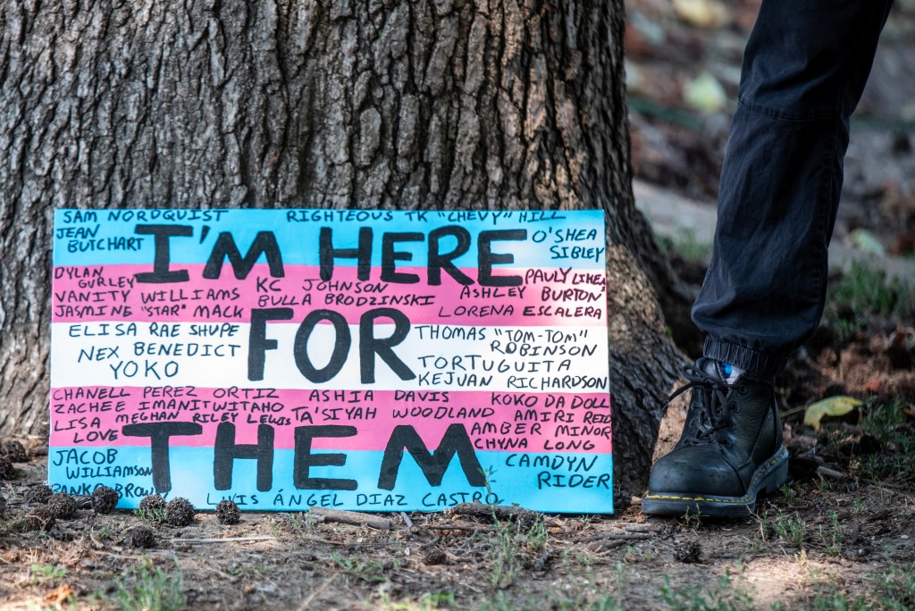 A close-up of a colorful sign propped against a tree, with the text 'I'M HERE FOR THEM' prominently displayed. The sign lists names of individuals, indicating solidarity with marginalized communities during a protest.