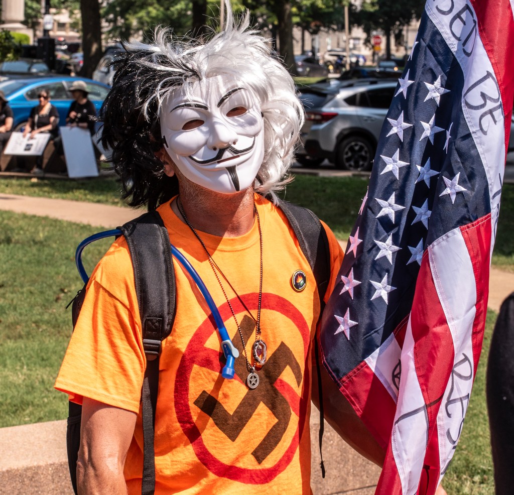 A person wearing a Guy Fawkes mask and an orange t-shirt with a symbol, holding an American flag at a protest.