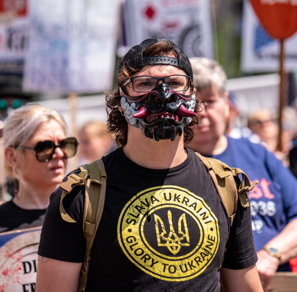 A protester wearing a decorative mask and a t-shirt that says 'SLAVA UKRAINI! GLORY TO UKRAINE!' stands in a crowd with signs in the background.