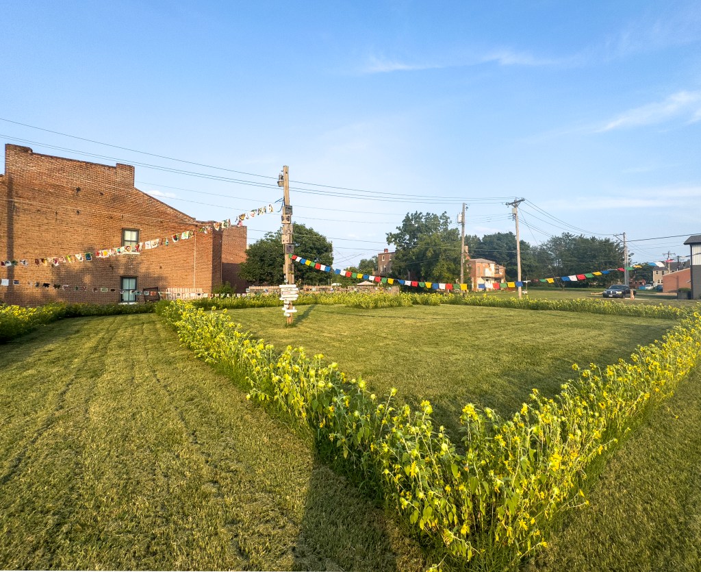 A green lawn area bordered by sunflowers, with colorful flags strung across two poles and a red brick building in the background under a clear blue sky.