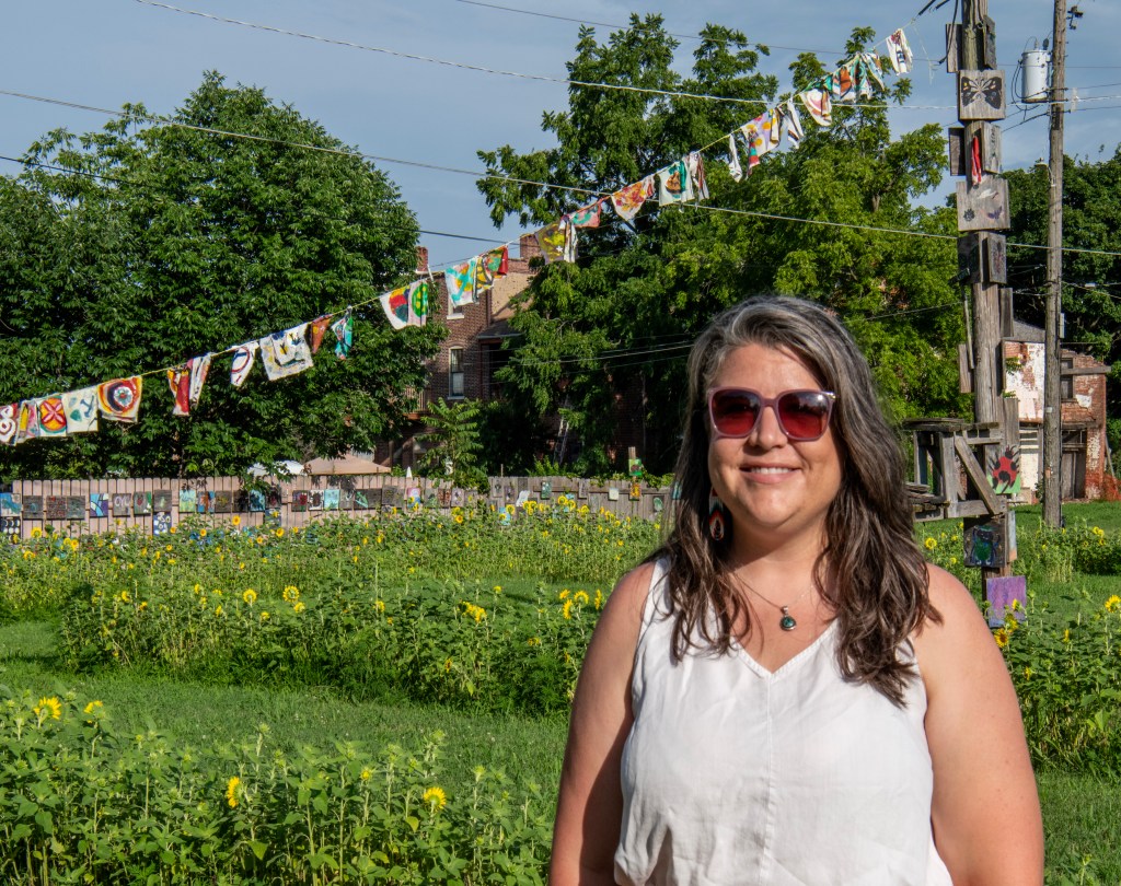 A woman with long hair and sunglasses stands in front of a field of sunflowers, with colorful fabric banners hung above in a community garden setting.