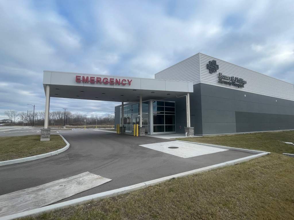 Exterior view of the emergency entrance to a hospital, featuring a gray building with a prominent 'EMERGENCY' sign and the hospital's name displayed on the facade.