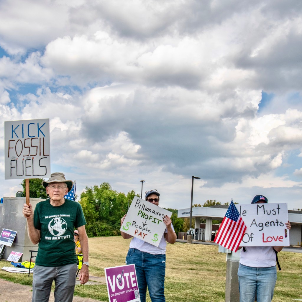 Two protestors holding signs at a demonstration. One sign reads 'KICK FOSSIL FUELS' and the other states 'Billionaires PROFIT vs People PAY'. The second protestor holds a small American flag. The background features a grassy area and overcast skies.