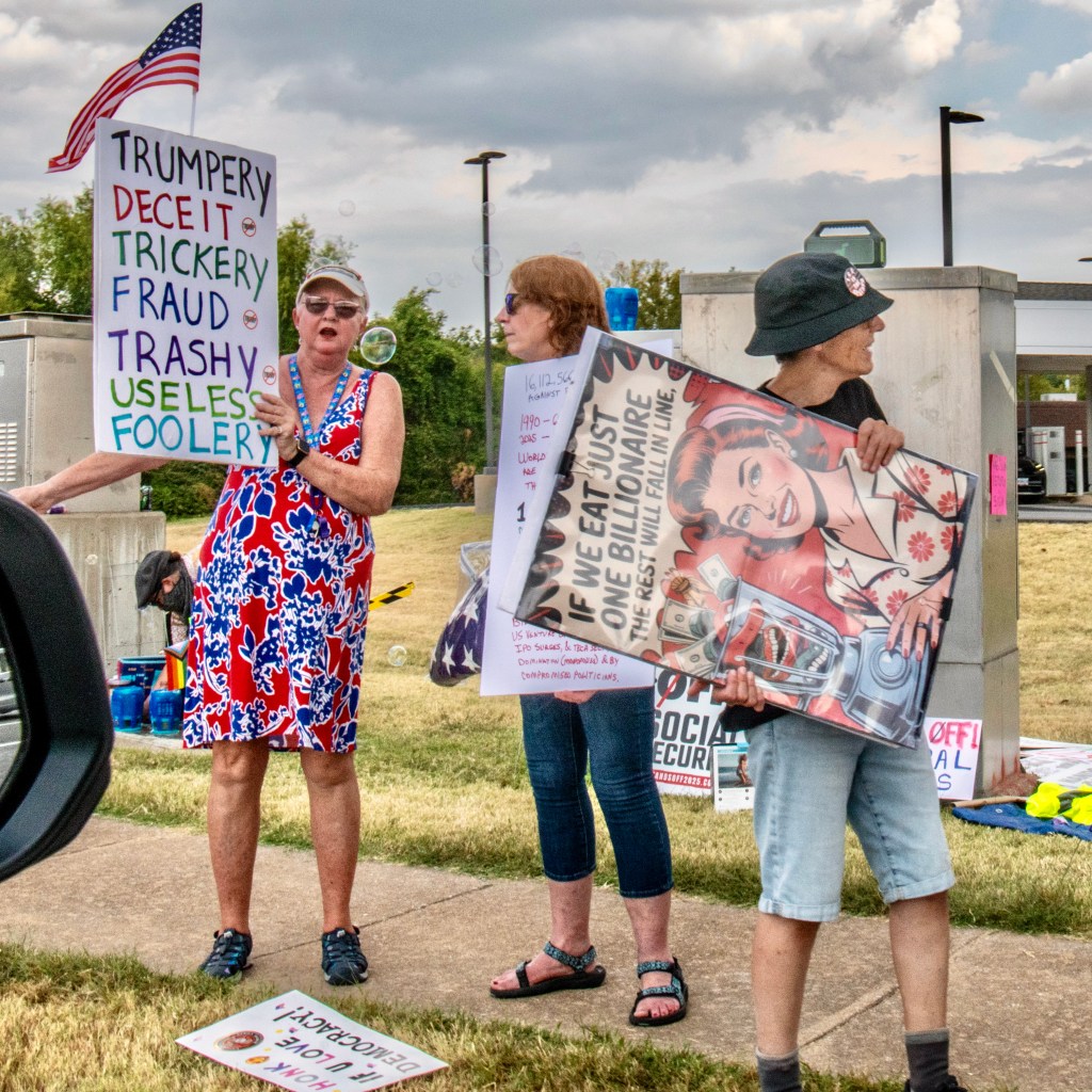 Three women participate in a protest holding signs with messages criticizing political issues and economic inequality, while an American flag is visible in the background.