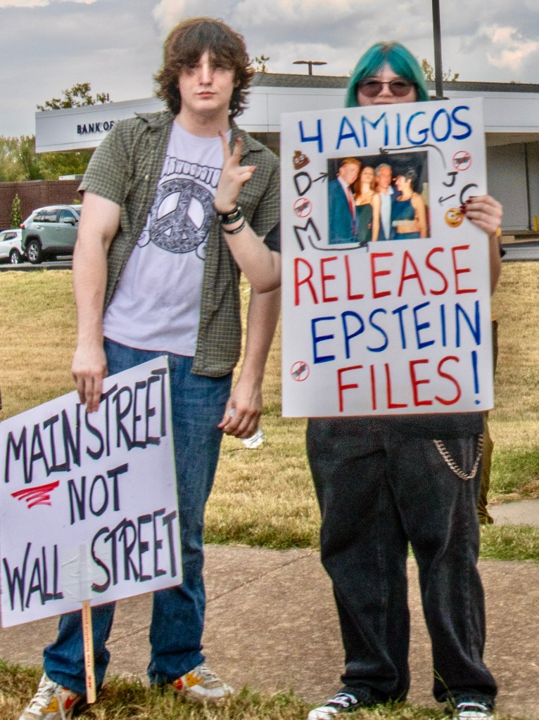Two protesters holding signs at a demonstration; one sign reads 'MAIN STREET NOT WALL STREET,' and the other says '4 AMIGOS RELEASE EPSTEIN FILES!'