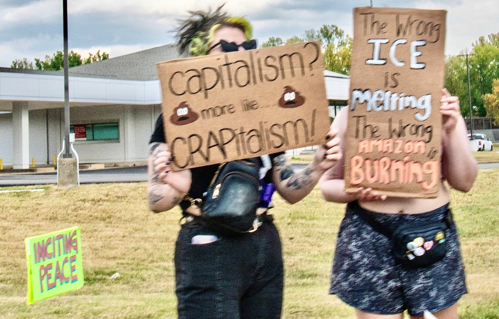 Two individuals hold up protest signs with messages criticizing capitalism and environmental issues during a rally.
