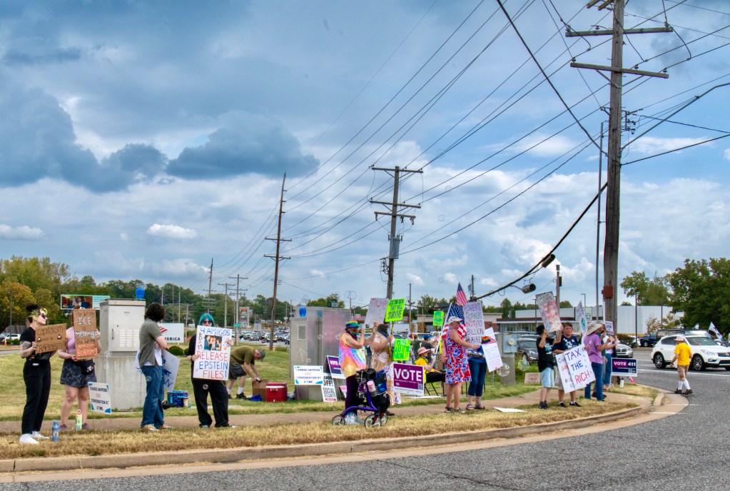 A group of protesters holding signs and flags at an event advocating for economic equality, located along Manchester Ave. in Ballwin, Missouri, under a cloudy sky.