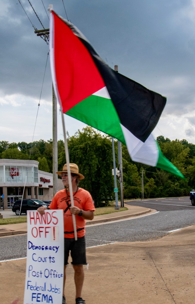 A protestor holding a Palestinian flag and a sign that reads 'HANDS OFF! Democracy Courts Post Office Federal Jobs FEMA' during a rally in Ballwin, St. Louis.