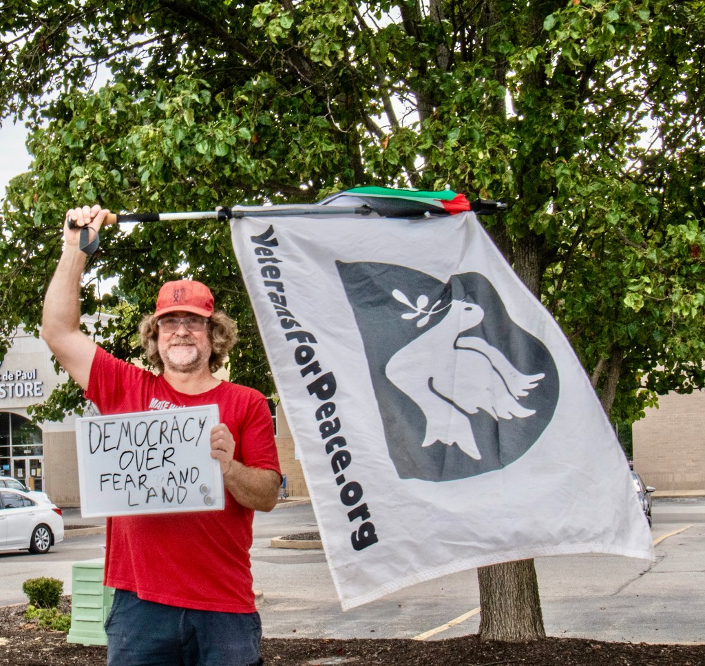 A man holding a sign that reads 'Democracy over fear and land' while standing under a tree, with a 'Veterans for Peace' flag waving beside him.