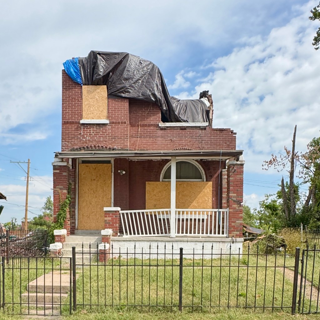 A damaged brick house with boarded-up windows, partially covered by a black tarp, amidst an overgrown grassy area.