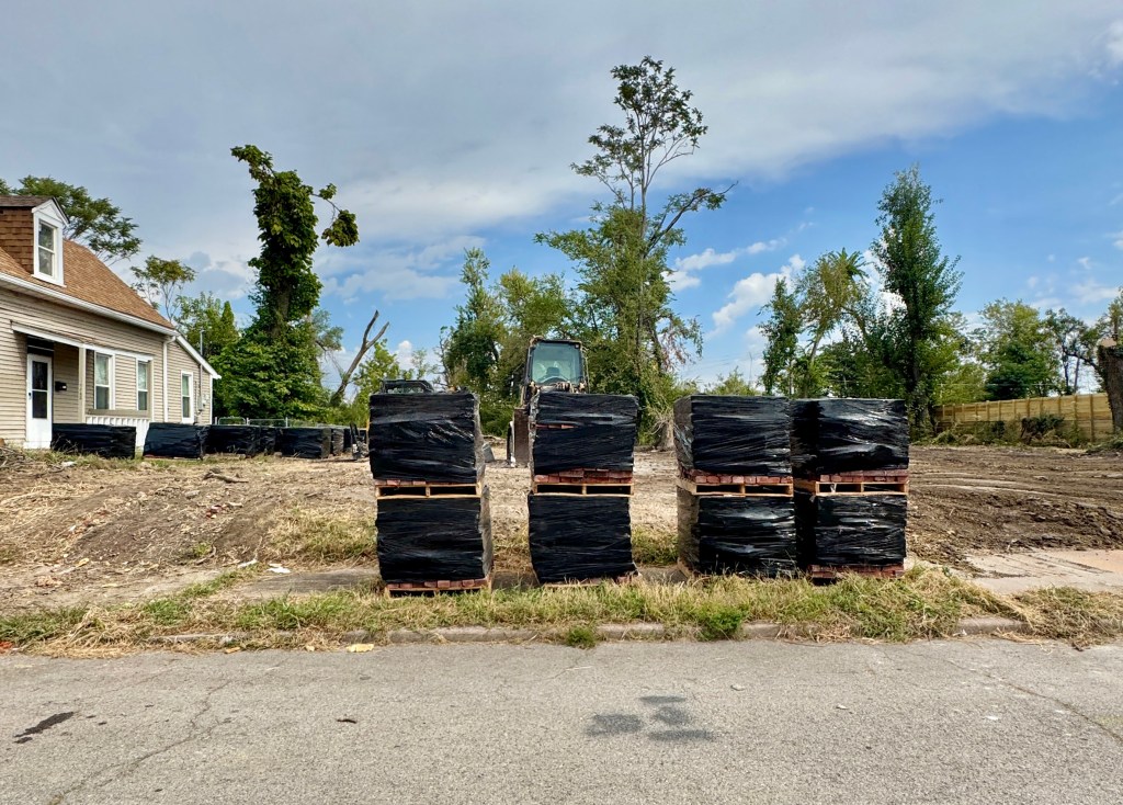 Pallets of materials wrapped in black plastic are stacked near an empty lot with a construction vehicle in the background, alongside a house and trees.