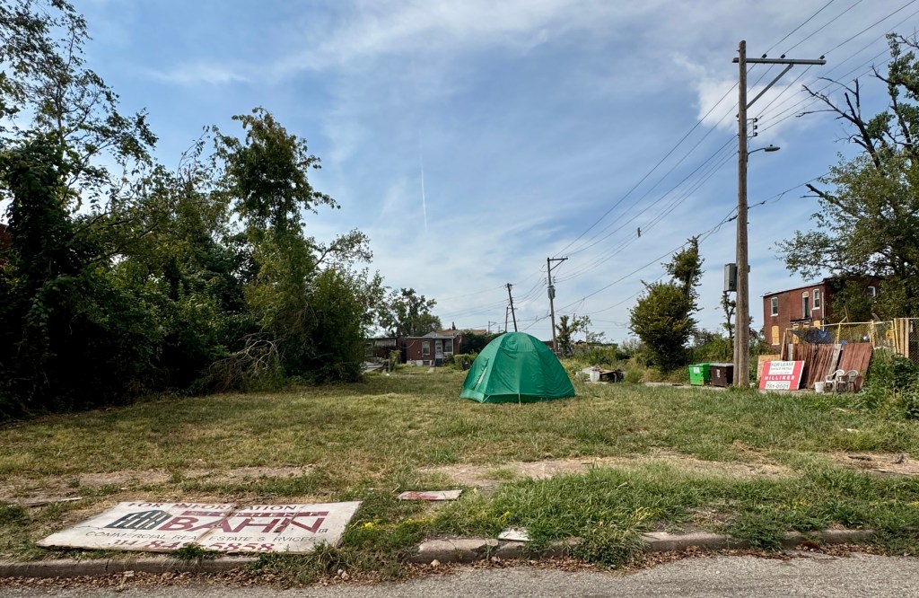 An empty lot in St. Louis with a green tent, overgrown grass, and nearby utility poles, showcasing the aftermath of urban displacement.