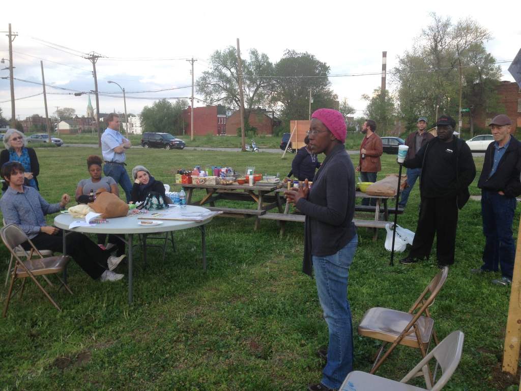 A community meeting is taking place outdoors with several people gathered around a picnic table, discussing and listening attentively. A woman stands in the foreground, speaking while others observe from their seats.