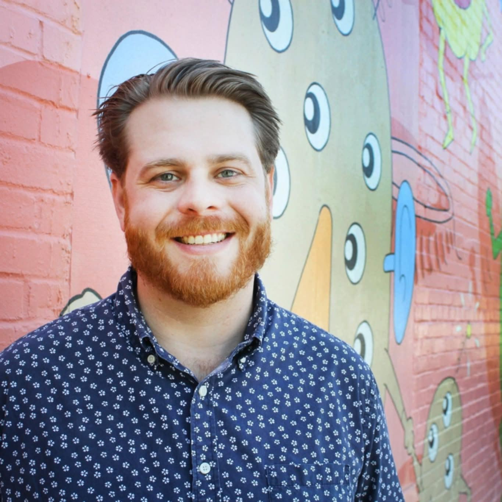 A smiling man with a beard stands in front of a colorful mural featuring whimsical characters and shapes. He wears a blue patterned shirt and appears cheerful.
