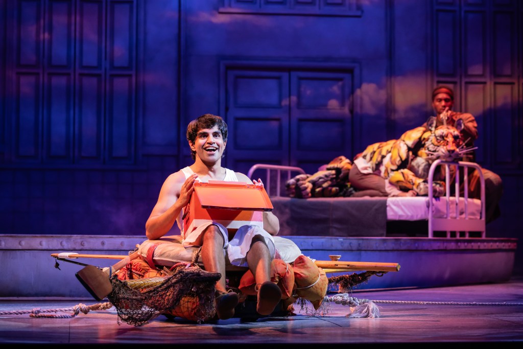 A scene from the play 'Life of Pi' showing Pi Patel sitting on a makeshift raft, smiling and holding a box, while a tiger puppet is resting on a hospital bed in the background.