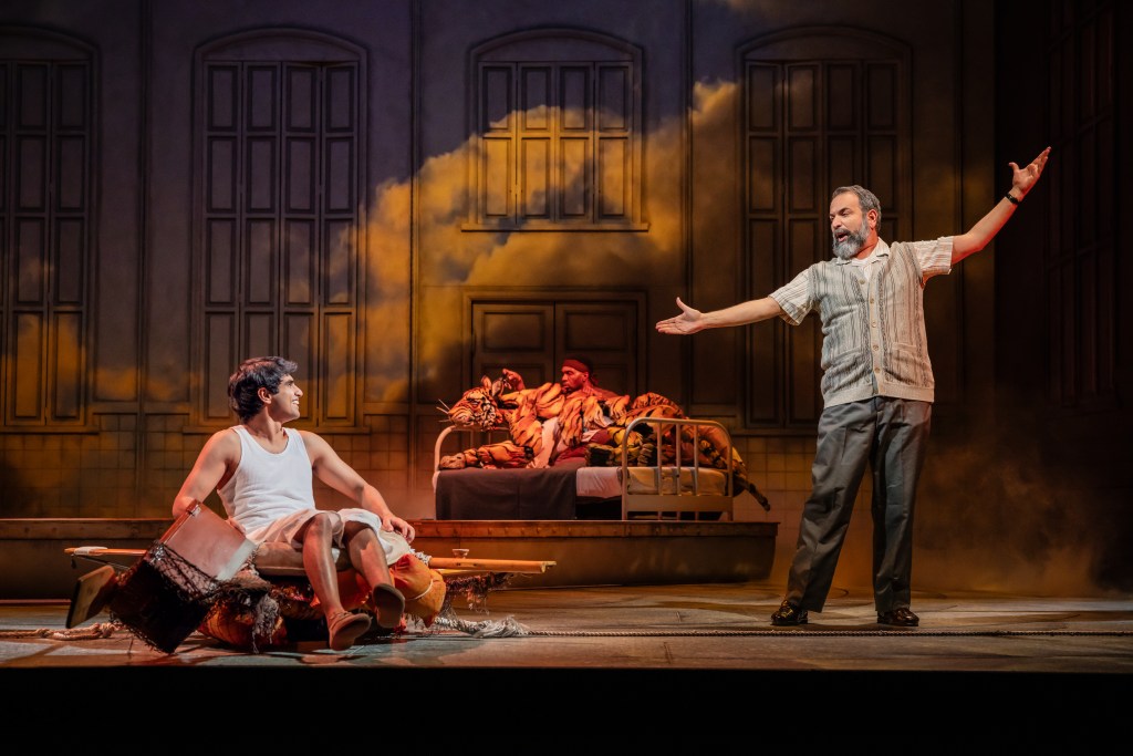 A scene from the play 'Life of Pi' featuring a teenage boy sitting on a raft, with a hospital bed and characters in the background, depicting a moment of storytelling and emotional interaction.
