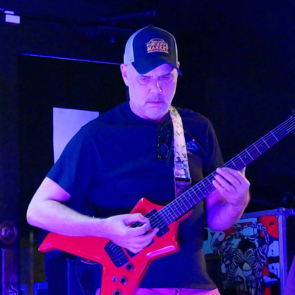 A musician playing a red electric guitar on stage under purple lighting.
