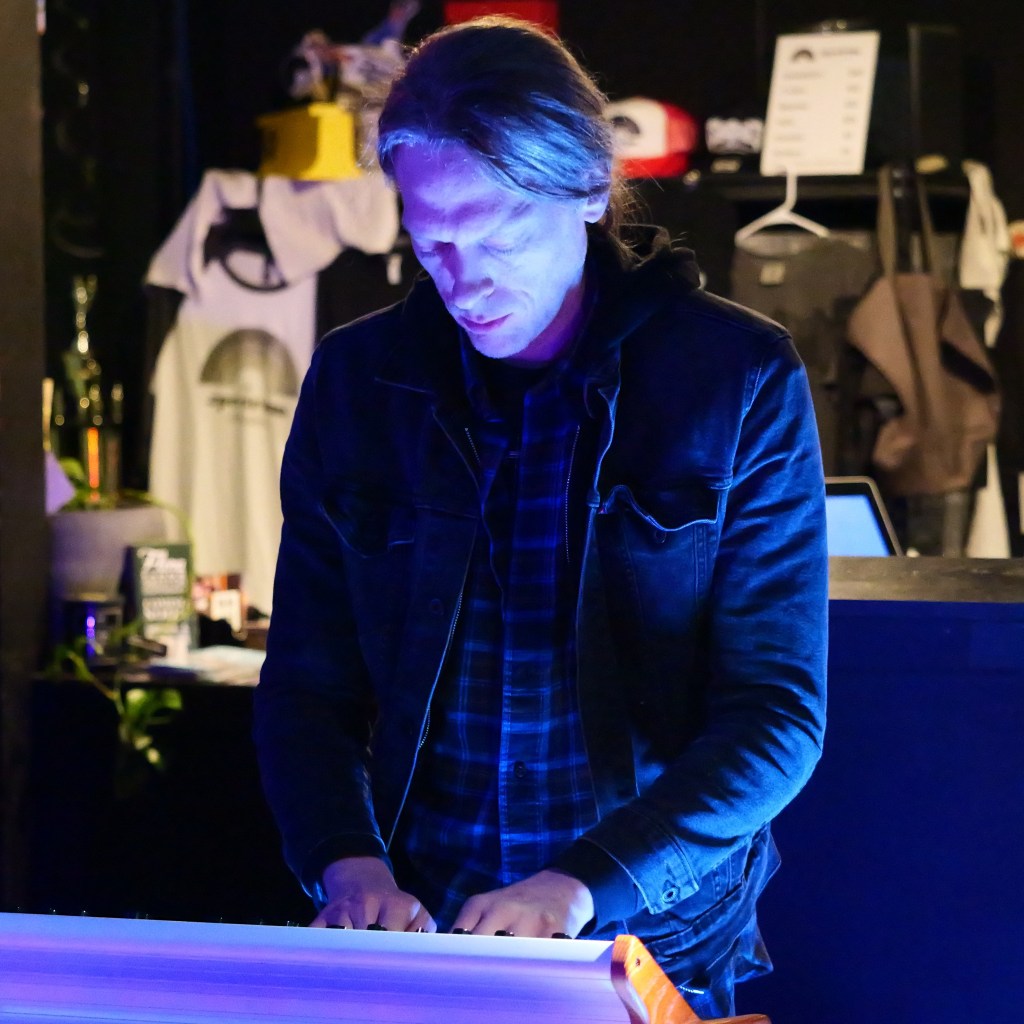 A person with long hair, wearing a denim jacket and plaid shirt, is playing a keyboard under blue lighting at a live music venue.