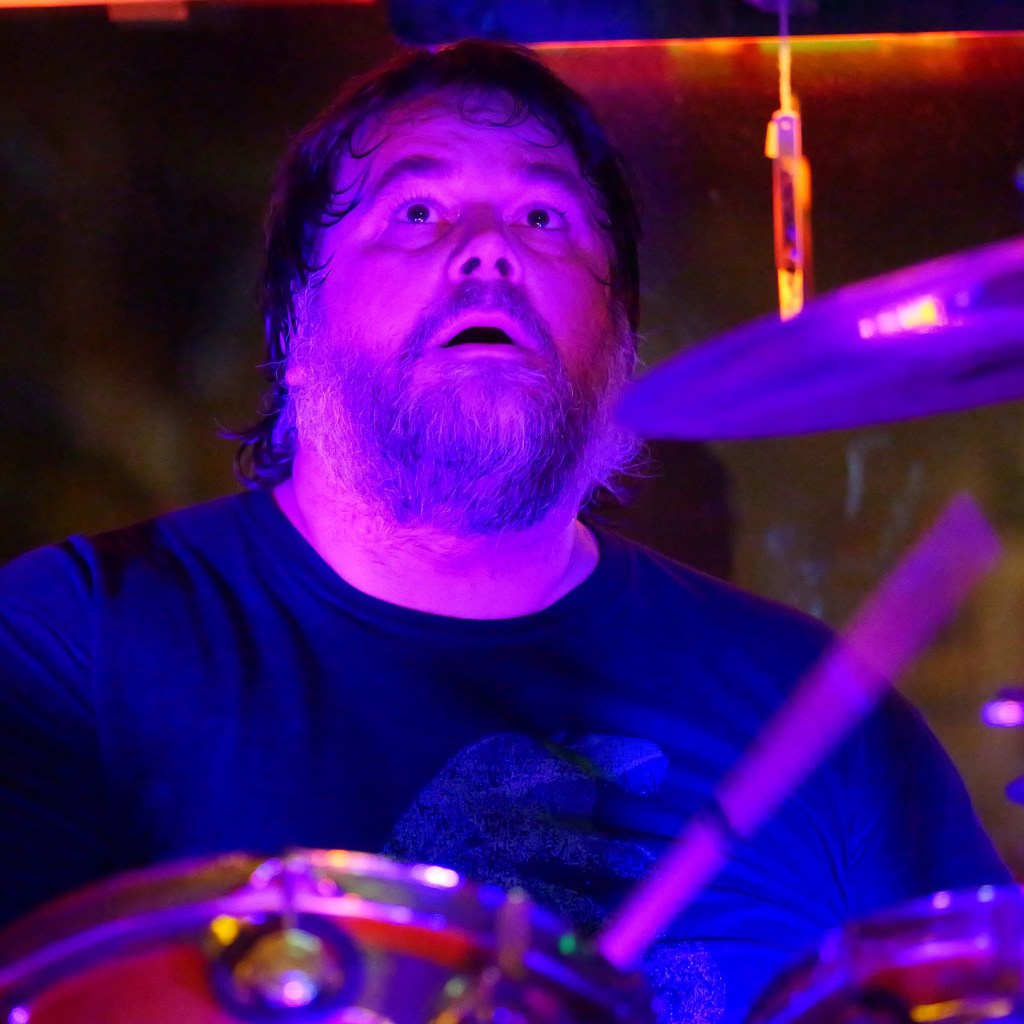 A drummer performing on stage, with a surprised expression, illuminated by purple stage lights.