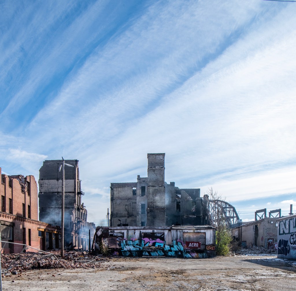 Ruins of the Crunden-Martin Manufacturing Company warehouses, with burnt debris scattered on the ground and smoke visible. The remains of the buildings feature exposed brick walls and graffiti on the standing structures, under a blue sky with wispy clouds.