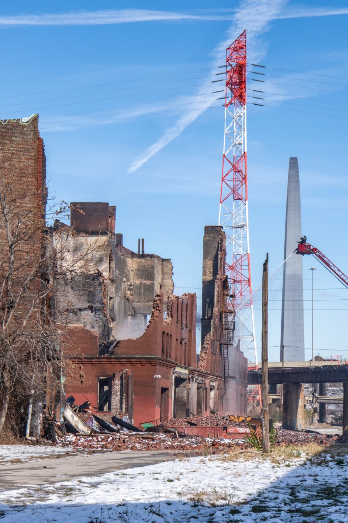 View of the charred and crumbling remains of the Crunden-Martin Manufacturing Company warehouses after a devastating fire, with a telecommunications tower and arch structure in the background.