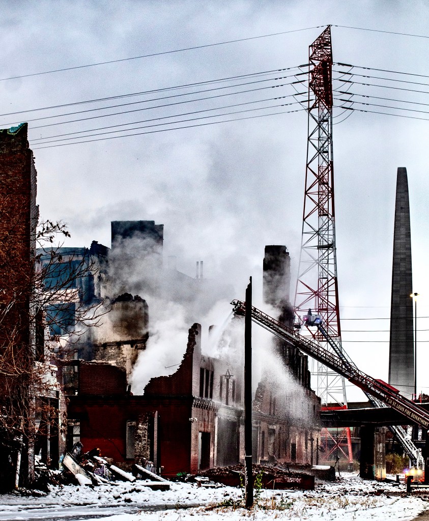 A fire response team spraying water onto the remains of a historic building engulfed in smoke, with electrical towers in the background.