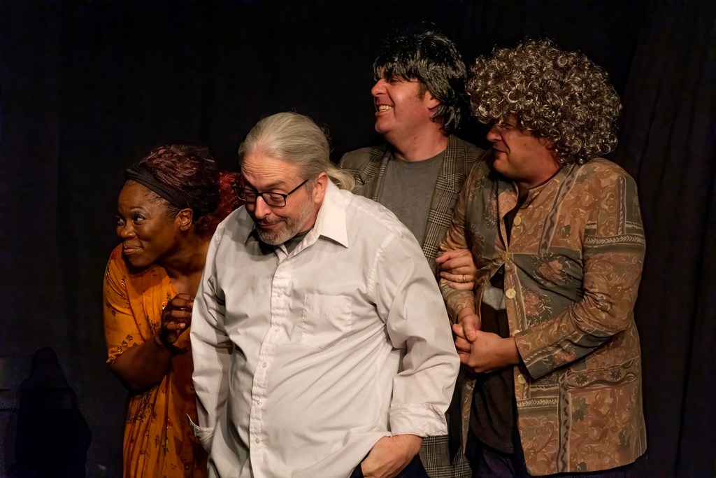 Four actors performing on stage, wearing various costumes and hairstyles, with a dark background.