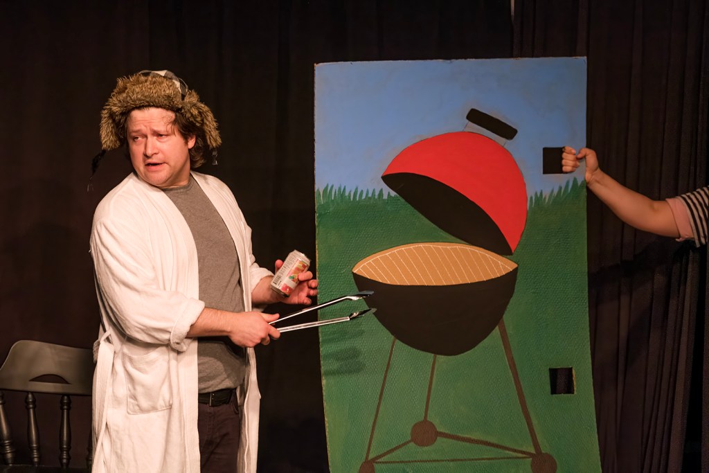A man wearing a bathrobe and a furry hat holds tongs and a can while standing next to a painted backdrop of a grill, suggesting a comedic performance.